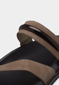 Καφέ σουέντ σανδάλι με δύο φαρδιές λουράκια, το ένα με γυαλιστερή μαύρη λεπτομέρεια, και μαύρο πέλμα με ορατή ραφή.