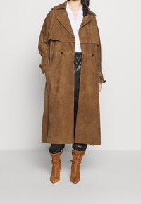 Trench coat in suede marrone con ampio colletto, design doppiopetto, due tasche frontali e vestibilità rilassata indossato sopra pantaloni in pelle nera e stivali marroni.