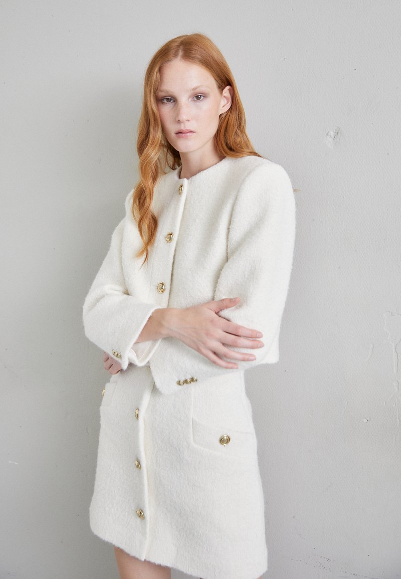 Λευκό σακάκι από μείγμα μαλλιού με χρυσά κουμπιά, με στρογγυλή λαιμόκοψη, μακριά μανίκια και μπροστινές τσέπες, απεικονιζόμενο σε ουδέτερο φόντο.