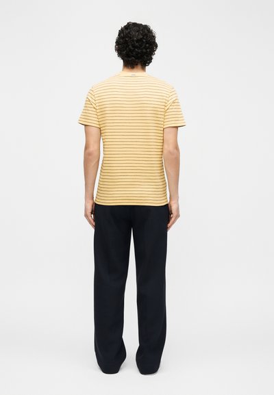 Άνδρας με σγουρά μαύρα μαλλιά που φοράει κοντομάνικο κίτρινο πουκάμισο με λεπτές οριζόντιες ρίγες και μαύρο χαλαρό παντελόνι, γυρισμένος από πίσω.