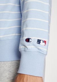 Světle modrá mikina se pruhy a bílými liniemi; žebrované manžety; na rukávu vyšívané loga Champion a MLB; měkká textura.