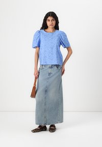 Haut bleu clair à manches bouffantes avec un motif floral, associé à une longue jupe en denim. Un sac à main marron et des chaussures à motif complètent la tenue.
