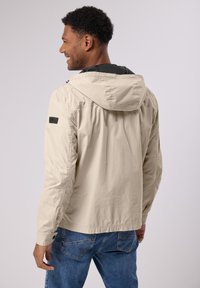 Beige lehká bunda s kapucí, se černou podšívkou a nášivkou na levém rukávu. Casual střih s hladkým povrchem.