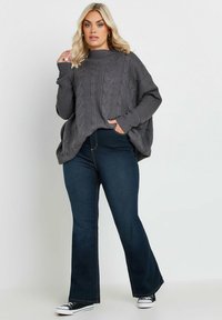 Grå stickad tröja med flätad design, hög hals och lös passform, tillsammans med mörkblå utsvängda jeans och svarta kanvas-skor.