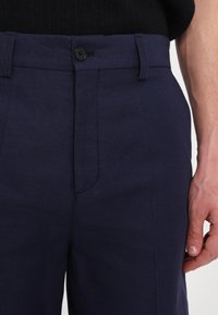 Pantalon bleu marine en tissu texturé, doté d'une fermeture à boutons sur le devant et de poches cousues, avec une coupe slim effilée.