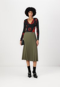 Top nero a maniche lunghe con motivo floreale rosso; gonna midi plissettata verde oliva; borsa nera; calzini neri alla caviglia; scarpe con tacco nere con fibbie.