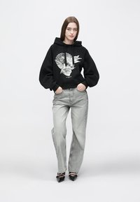 Must üle mõõdu suured kapuutsiga dressipluusid, millel on graafiline kolju kujundus, kombineeritud helehallide laiade pükstega ja mustade teravate kontsadega kingadega.