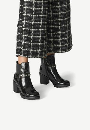 Bottines en cuir verni noir avec un talon épais, présentant une ceinture décorative et des accents cloutés. Stylisées avec une jupe à carreaux noir et blanc.
