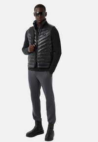 Zwarte gewatteerde bodywarmer over een donker truitje, gecombineerd met grijze broek en zwarte enkellaarsjes. Geaccentueerd met oversized zonnebril en een tas.