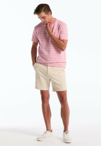 Polo shirt rayé rose clair avec un col rond, short beige et baskets blanches. La chemise présente un petit logo sur la poitrine.