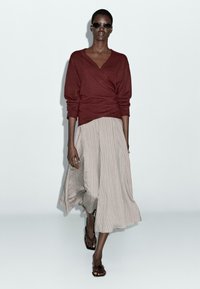 Burgundy knitted wrap sweater, beige striped midi skirt, model wearing dark sunglasses and brown flat sandals, textured fabric details.