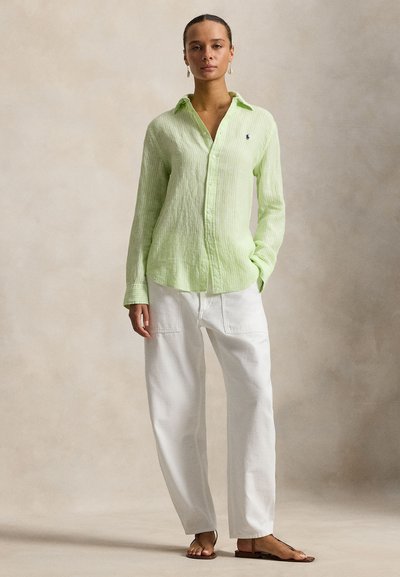 Λαχανί πουκάμισο με μακριά μανίκια και κάθετη υφή, σε συνδυασμό με λευκό παντελόνι φαρδιάς γραμμής και καφέ επίπεδα σανδάλια. Μινιμαλιστικός σχεδιασμός, χωρίς σχέδια.