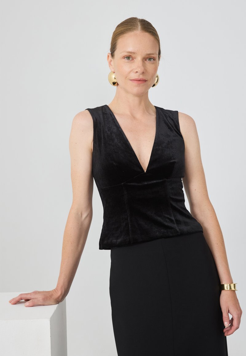 Top sin mangas de terciopelo negro con un escote en V profundo, que cuenta con pinzas estructuradas en la cintura. Una falda negra y una pulsera dorada en el brazo completan el look.