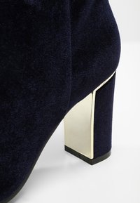 Zapato de tacón alto de terciopelo azul marino con un tacón grueso acentuado en dorado y una textura suave, que resalta un diseño moderno.