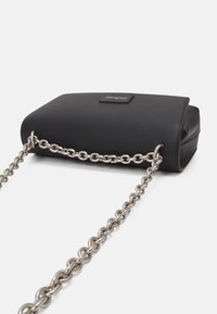 Svart handväska med räfflad textur, rektangulär form och en silverkedja som rem. Har en varumärkeslogotyp på en rektangulär tagg.