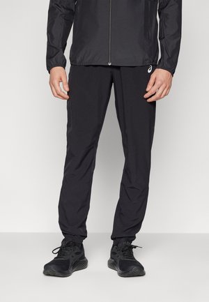 Hombre con chaqueta deportiva negra, pantalones de chándal negros con logo y zapatillas de correr negras, de pie sobre un fondo liso.