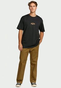 Jovem em pé com uma mão no bolso, usando uma t-shirt preta estampada, calças castanhas e tênis preto e branco, em fundo liso.
