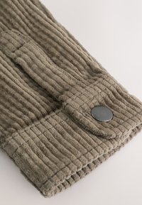 Tissu en velours côtelé beige avec des rainures verticales, orné d'un bouton noir et de surpiqûres. Gros plan sur un poignet de manche.