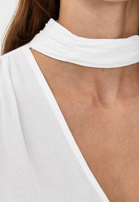 Vit topp med draperad krage, tillverkad av ett mjukt, texturerat tyg. Designen inkluderar en djup V-ringning och subtila sömnadsdetaljer.