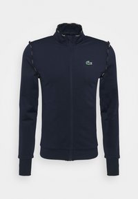 Mörkblå zip-up jacka i mjukt material med hög krage, sidofickor och en liten grön Lacoste-logotyp på bröstet.