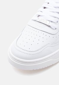 Κοντινό πλάνο άκρου λευκού δερμάτινου αθλητικού παπουτσιού με διατρήσεις, λευκά κορδόνια και μεταλλική ετικέτα κορδονιών χαρ αγμένη με το όνομα "Steve Madden".