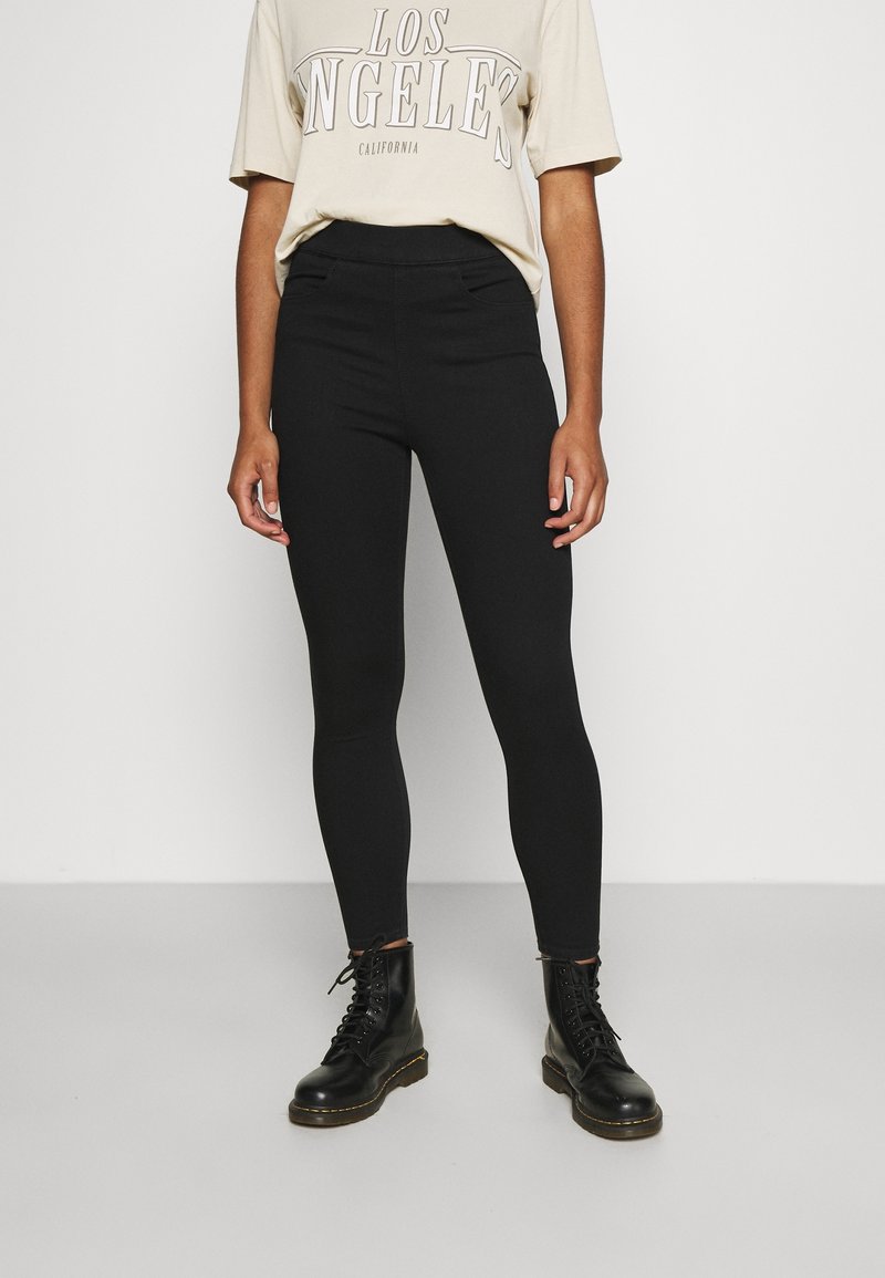 Personne portant un t-shirt beige "Los Angeles California", des leggings noirs taille haute et des bottes noires à lacets, debout devant un fond uni.