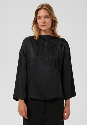 Vrouw met blond haar die een loszittende zwarte top met lange mouwen en franjes op één schouder draagt, staand tegen een grijze achtergrond.