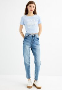 Camiseta corta de color azul claro con un gráfico que dice "SURF CAMP", combinada con unos vaqueros de tiro alto de color azul deslavado y zapatillas blancas con suela color marrón.