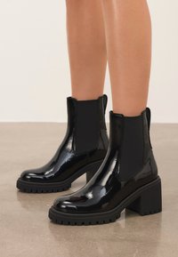 Shiny black ankle boots with chunky heels and elastic side panels worn on bare legs, standing on a smooth polished floor.