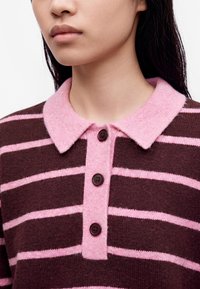 Gros plan d'une personne portant un pull rayé marron et rose avec un col rose et trois boutons foncés sur le devant.