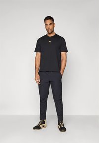 Zwart T-shirt met korte mouwen en een gouden embleem, gecombineerd met zwarte taps toelopende broek en zwart-beige sportschoenen.