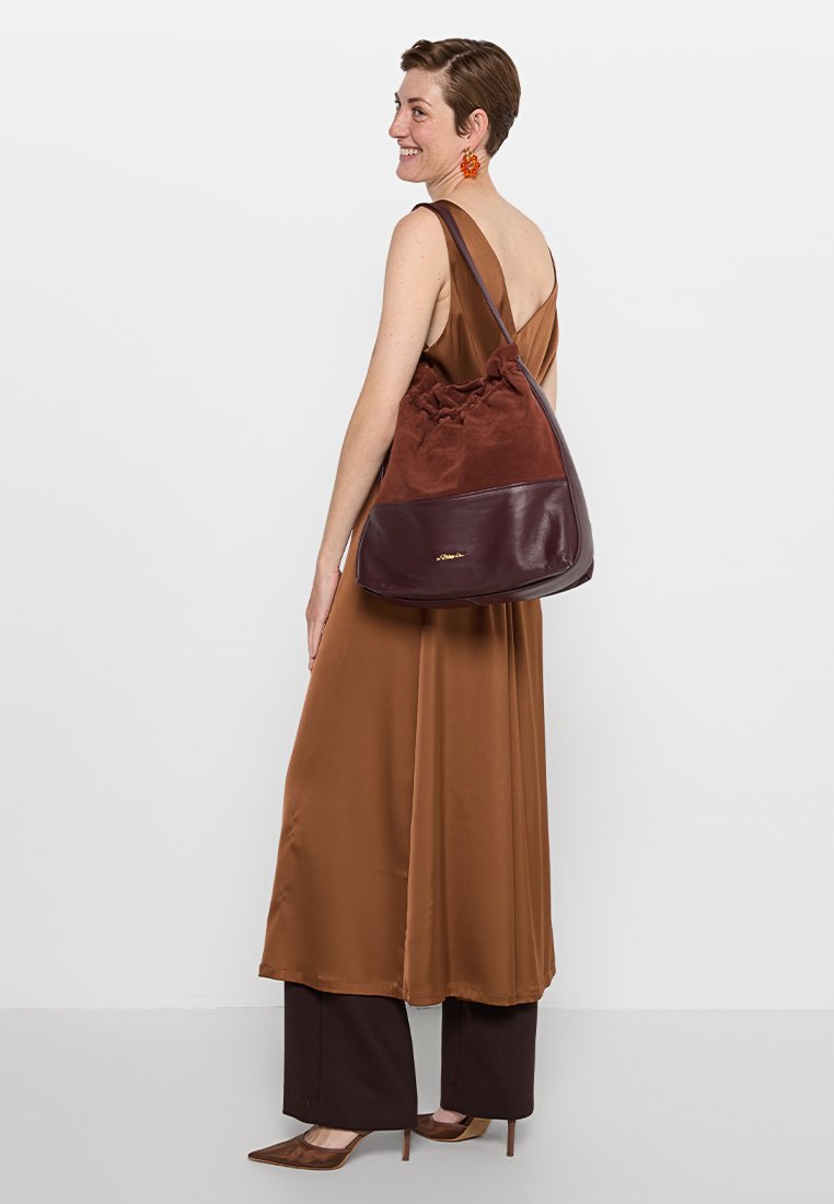 Kobieta uśmiechająca się, ubrana w brązową sukienkę bez rękawów i spodnie, trzymająca dużą torebkę na ramię w dwu-tonowym brązowym skórzanym i zamszowym stylu.
