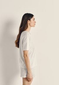 Žena s dlhými hnedými vlasmi stojaca v profile, oblečená v bielej košeli s krátkym rukávom a svetlosivých šortkách na jednoliatom béžovom pozadí.