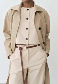 Beige trenčkot přes světle béžový top s černými knoflíky, spárovaný s vysokými béžovými kalhotami a hnědým páskem se zlatou kulatou sponou.