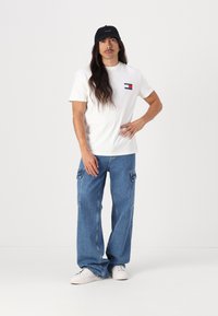 Hvit bomulls t-skjorte med et lite logo-patch, sammen med løse blå denim cargo-bukser og hvite joggesko, akkompagnert av en svart caps.
