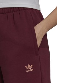 Bordeauxfärgade joggingbyxor i mjukt tyg, med sidofickor och en broderad ljusrosa Adidas-logga på nederbenet. Elastisk midja.