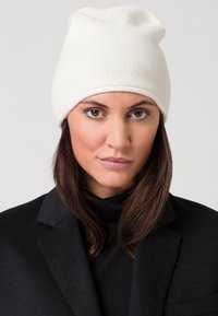 Berretto in maglia bianco con una texture morbida, design leggermente scivolato. Indossato con un cappotto nero, evidenziando il contrasto dei colori.