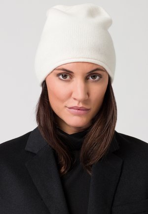 Berretto in maglia bianco con una texture morbida, design leggermente scivolato. Indossato con un cappotto nero, evidenziando il contrasto dei colori.
