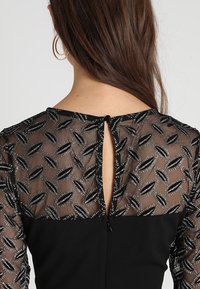 Svart klänning med en genomskinlig, broderad nätbakgrund med ett bladmönster. Nyckelhålsöppning med knappstängning vid halsringningen.