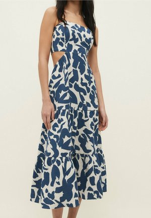 Vrouw draagt een mouwloze midi-jurk met blauw-wit patroon en zijopeningen en een gelaagde rok, staand voor een effen achtergrond.