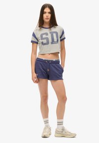 Grijze cropped t-shirt met marineblauwe strepen en "SD" opdruk, gecombineerd met marineblauwe shorts en lichte sneakers. Voorzien van een trekkoord in de taille.