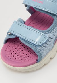 Sandale bleu clair avec semelle intérieure rembourrée rose, équipée de deux sangles ajustables en Velcro et d'un upper en maille respirante. Semelle extérieure texturée pour une meilleure adhérence.