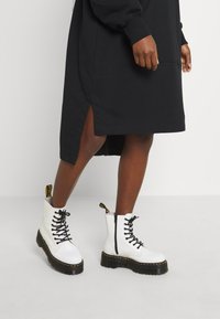 Weiße Knöchelstiefel mit schwarzen Schnürsenkeln, seitlichem Reißverschluss und gelbem Schlaufen. Schwarzes Kleid mit sichtbaren Seiten-Schlitzen, getragen an einem menschlichen Bein.