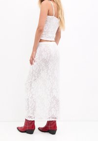 Top corto de encaje blanco combinado con una falda larga de encaje. Presenta tirantes finos y un patrón floral. Se lleva con botas de vaquero rojas brillantes.