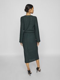 Πλεκτό φόρεμα σε �σκούρο πράσινο χρώμα με μακριά μανίκια, μήκος ως το μέσο της γάμπας, υφή σαν δαντέλα και εφαρμοστή γραμμή. Φορεμένο με μαύρες ψηλοτάκουνες γόβες.