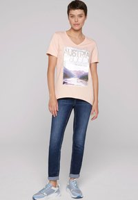 Jasnoróżowy t-shirt z dekoltem w serek z grafiką góry i napisem. W połączeniu z ciemnoniebieskimi dżinsami i szaro-niebieskimi butami sportowymi.