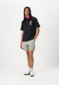 Sort t-shirt med korte ærmer og farverig grafik, lysegrønne shorts og hvide sneakers. Modellen har et rødt bandana om halsen.