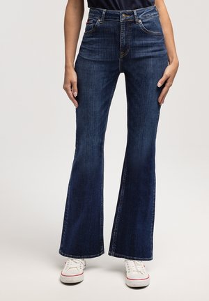 Kobieta ubrana w ciemnoniebieskie dzwony i białe sneakersy stoi na tle jasnym.