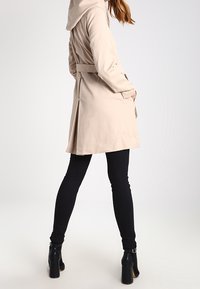 Beige trench coat with a hood, belted waist, and side slits, paired with black skinny jeans and high-heeled black boots.