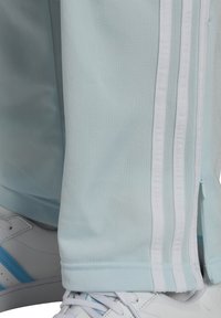 Světle modré sportovní kalhoty s bílými bočními pruhy, vyrobené z hladkého materiálu, s kapsou na zip a zúženým střihem u kotníků.